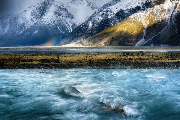 By Michael Piraino River flowing in front of mountains and small rainbow