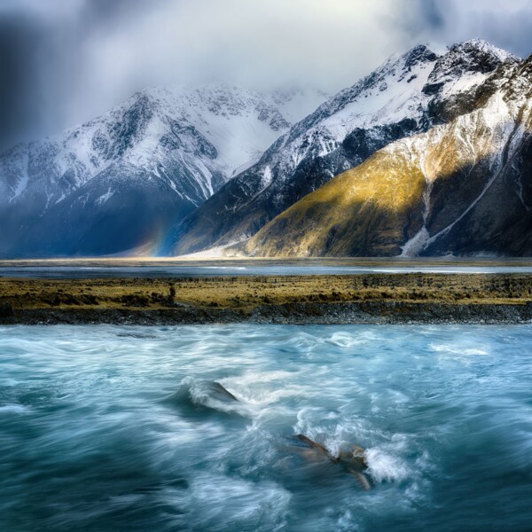 By Michael Piraino Flowing water in the foreground with small rainbow and mountains in the distance, New Zealand